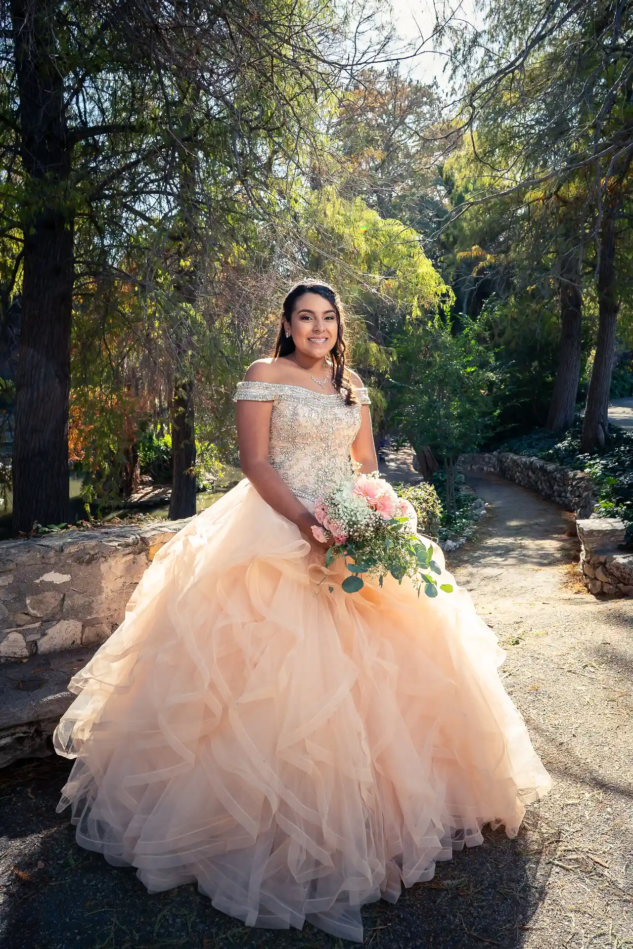 Quinceañera in a peach ruffled gown in a garden with trees and a stone wall