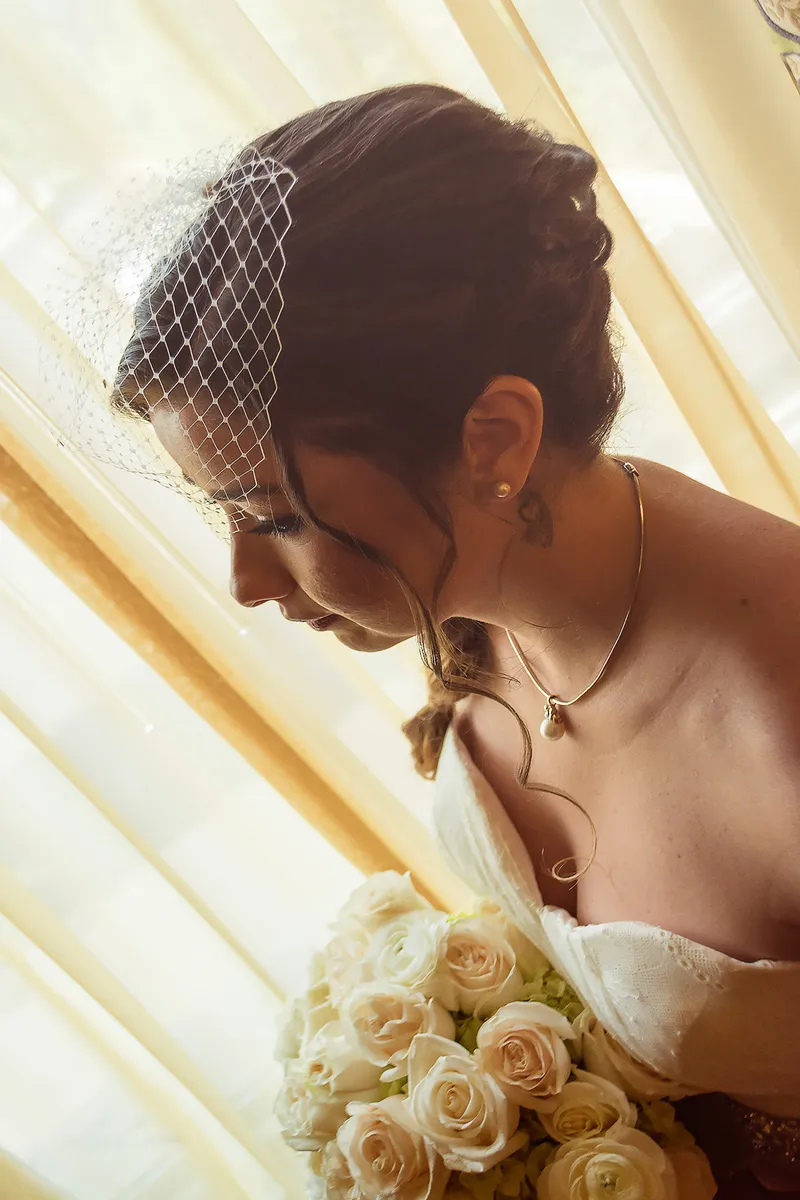 Bride in profile wearing a birdcage veil and holding her bouquet in soft window light