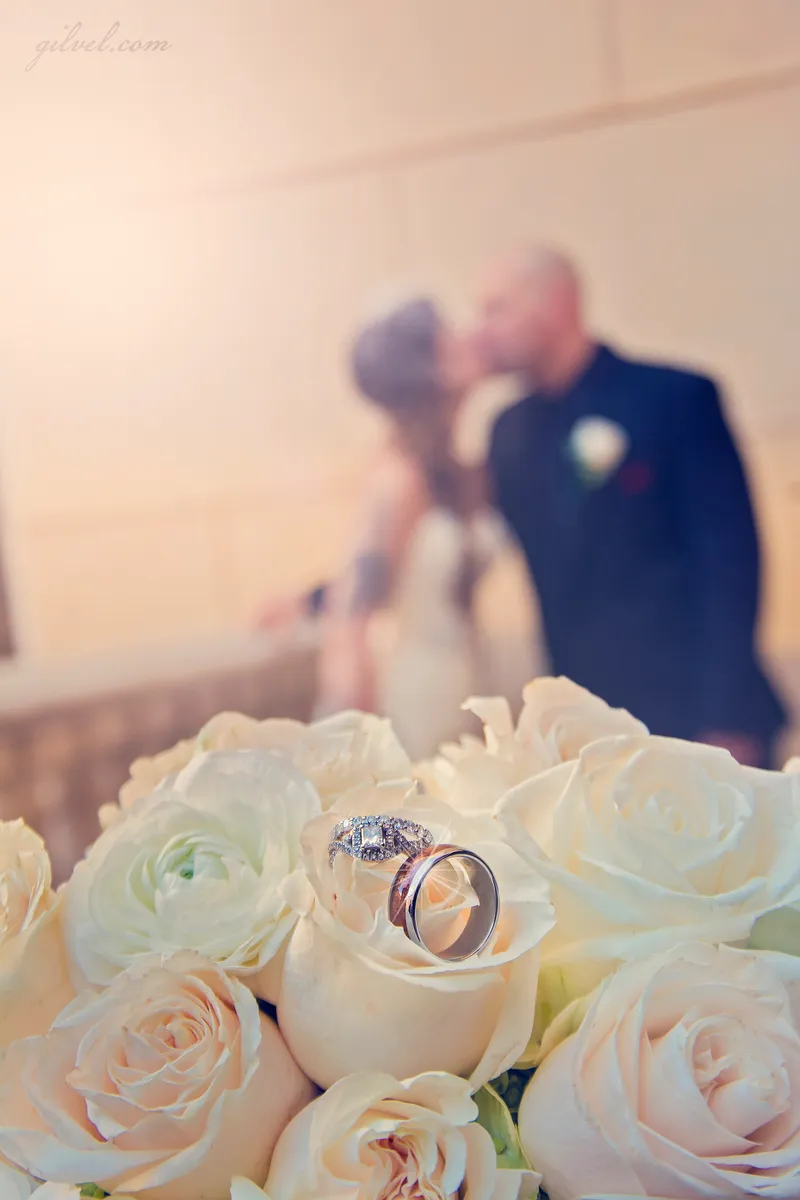 Two rings resting on a white rose bouquet with a couple kissing, blurred, in the background